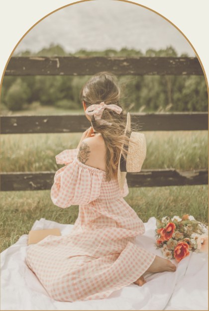 A girl in a pink gingham dress sitting on a blanket by a fence with a basket of roses